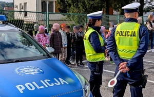 Umundurowana policjantka i policjant ruchu drogowego, po lewej radiowóz, a tle grupa dzieci.