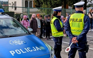 Umundurowana policjantka i policjant ruchu drogowego, po lewej radiowóz, a tle  grupa dzieci.