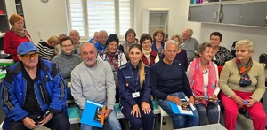 Grupa seniorów siedzących na ławkach, po środku umundurowana policjantka