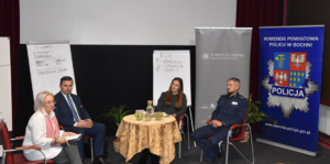 Umunduorwany policjant oraz osoby prowadzące spotkanie siedzące na krzesłach obok okrągłego stolika. Na zdjęciu widoczne banery Komendy Powiatowej Policji w Bochni oraz Starostwa Powiatowego w Bochni