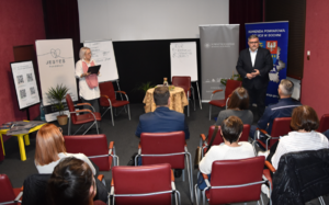 Starosta Bocheński stojący obok banerów Starostwa Powiatowego w Bochni oraz Komendy Powiatowej Policji w Bochni. Na zdjęciu widoczna osoba prowadząca spotkane oraz uczestnicy siedzący na krzesłach