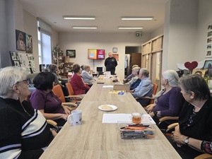 Seniorzy siedzący przy stole i dzielnicowy piotr prowadzący pogadankę