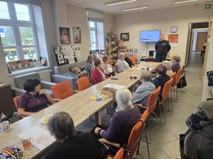 Seniorzy zgromadzeni przy stole i dzielnicowy przekazujący informacje