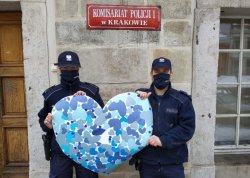 Policjantka i policjant stoją przed wejściem do budynku komisariatu policji pierwszego i wspólnie trzymają duże serce zrobione z wyciętych z papieru małych niebieskich serc