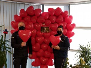 2 Komendant Komisariatu policji szóstego wraz z policjantką stoją w pomieszczeniu i trzymają w dłoniach balony w kształcie serca, za nimi duże serce w wykonane z czerwonych balonów