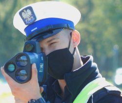 policjant ruchu drogowego (wyeksponowana czapka z białym pokrowcem) w ręce trzyma ręczny miernik prędkości koloru granatowego