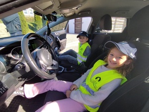 Dziewczynka i chłopiec w kamizelkach odblaskowych i policyjnych czapkach, siedzą w radiowozie