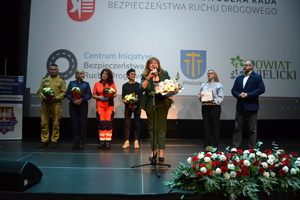 Pani Wicestarosta przemawia, Strażak, policjantka, pielęgniarka i Jakub uczestnik wypadku stoją na scenie trzymając w ręku kwiaty