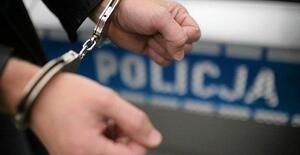 Na rękach założone są policyjne kajadanki. W tle fragment radiowozu z napsem policja