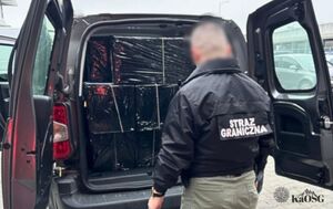 Funkcjonariusz Straży Granicznej w kurtce z napisem &amp;quot;STRAŻ GRANICZNA&amp;quot; stoi tyłem przy otwartym bagażniku samochodu dostawczego, który jest wypełniony zatrzymanymi, czarnymi paczkami.