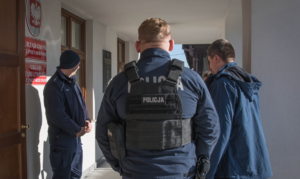 Umundurowani policjanci oraz osoba biorąca udział w ćwiczeniach. Na fasadzie budynku widoczne godło Polski oraz tablice z nazwaniu urzędów
