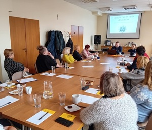 Przy stole siedzi policjant i policjantka, a wokoło osoby. W tle ekran z prezentacją.