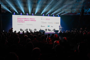 Tauron Arena, na hali mrok,  podświetla  się rzutnik, przy mównicy stoi mężczyzna