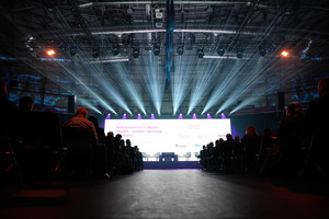 Ustawione rzędy krzeseł, na hali panuje mrok, podślwitlony rzutnik, na którym wyswietlony jest tekst, nad tekstem oświetlone jupitery