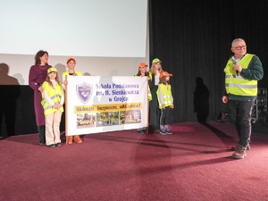 Na scenie dzieci wraz z nauczycielką trzymają w ręku plakat Szkoła Podstawowa w Grojcu, obok redaktor Koczyba trzymający w ręku mikrofon