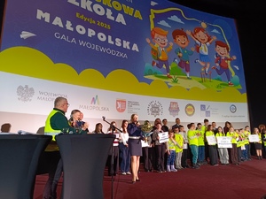 Na scenie na tle kolorowego ekranu przedstawiającego plakat Odblaskowej Szkoły stoją laureaci konkursu, wręczający nagrody raz przemawiająca policjantka