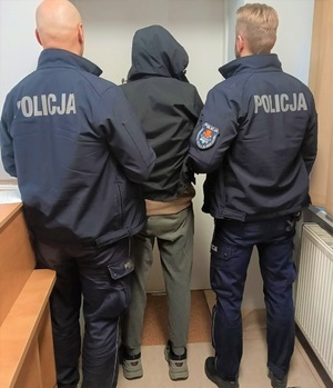 Dwóch umundurowanych policjantów, a w środku zatrzymany mężczyzna w kapturze