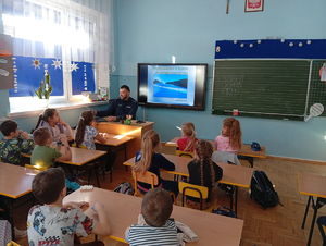 Policjant siedząc w klasie omwaia dzieciom wyświetlaną na tablicy prezentację o bezpieczeństwie w zimie.