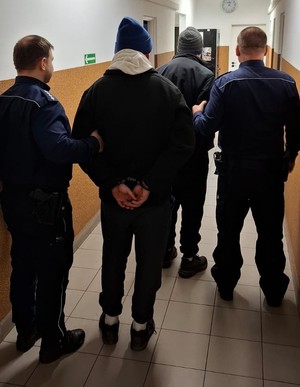 Policjanci stoją z zatrzymanymi mężczyznami. Mężczyźni mają nałożone kajdanki na ręce trzymane z tyłu.