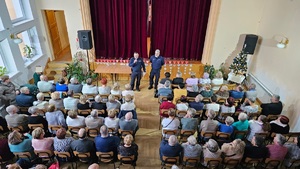 Na zdjęciu dwóch umundurowanych policjantów, a przed nimi na krzesłach siedzą osoby starsze.