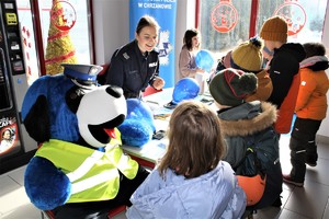 Policjantka stoi przy stoisku z materiałami profilaktycznymi, naprzeciwko stolików 6 dzieci stoi i uczestniczy w rozmowie