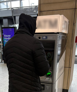 mężczyzna w czarnej kurtce z założonym kapturem stojący przed bankomatem w jednej z galerii handlowych - zdjęcie poglądowe