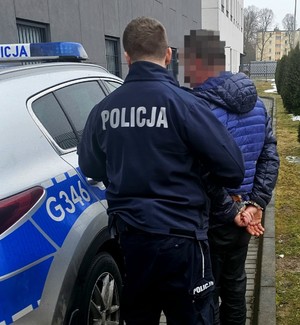 Na zdjęciu umundurowany policjant trzymający mężczyznę, który ma na rękach z tyłu kajdanki. Obok radiowóz