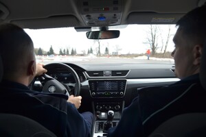 Dwóch mężczyzn siedzących w pojeździe, jeden z nich trzyma ręce na kierownicy