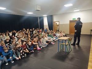 Siedzące na materacach dzieci ze szkoły podstawowej i policjant z drogówki stojący przed nimi