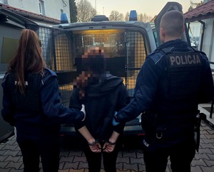 Policjant i policjantka, którzy trzymają zatrzymaną kobietę, która ma założone kajdanki na rękach z tyłu. W tle radiowóz