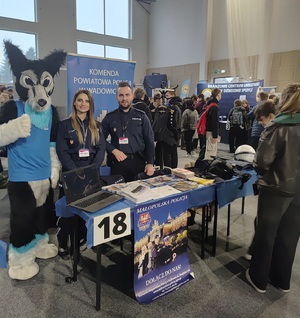 Przy stoliku policjant i policjantka oraz maskotka wilka. W tle banery. Na stole ulotki i sprzęt policyjny