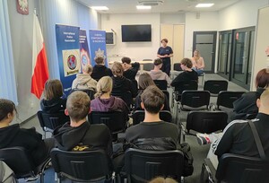 Uczniowie siedzący na krzesłach, przed nimi stoi policjantka i na krześle siedzi policjantka w stroju cywilnym