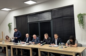 Przy stole siedzą dwie kobiety oraz czterech mężczyzn w strojach cywilnych oraz umundurowany policjant.