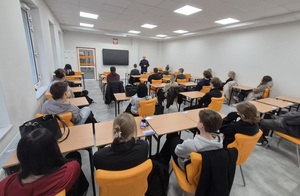 Uczniowie siedzący w szkolnych ławkach, a przed nimi policjant