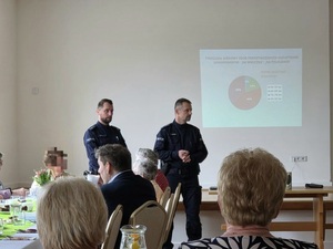 Dwóch policjantów, przed nimi osoby starsze siedzące przy stolikach. W tle wyświetlana prezetacja.