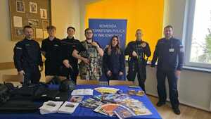 Na zdjęciu policjantka i dwóch policjantów oraz uczniowie klas mundurowych. W tle policyjny baner. Na stoliku sprzęt policyjny i ulotki