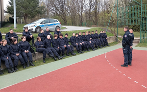 Umundurowani policjanci oraz uczniowie w mundurach policyjnych podczas prelekcji. Na zdjęciu widoczny policyjny radiowóz