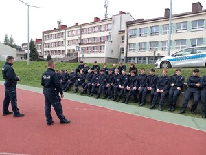 Budynek szkoły, policyjny radiowóz, policjanci oraz uczniowie w mundurach ćwiczebnych podczas prelekcji