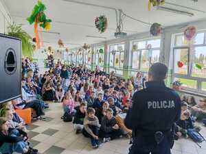 Policjant stoi tyłem. Przed nim na korytarzy szkołu siedzą uczniowie_