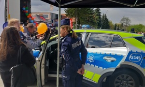 Policjantka i oznakowany radiowóz. W tle grupa osób