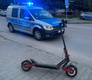 Na zdjęciu oznakowany radiowóz i hulajnoga elektryczna. W tle blok i samochody