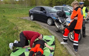 Bezpieczna majówka lekcja bezpieczeństwa kierowca uczeń i policjant w tle radiowozy i motocykl