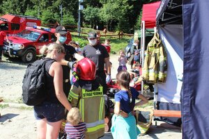 Strażacy tłumaczą dwóm dziewczynkom stojącym z mamą jak wygląda praca strażaka i jakim sprzętem się posługują w czasie pożaru. Jedna dziewczynka ma założony na siebie hełm strażacki