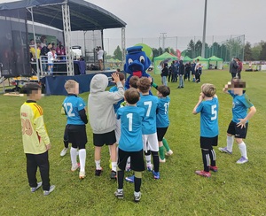 Na boisku maskotka Wawelka i grupa chłopców przybijająca z nim piątkę. W tle osoby