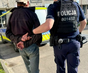 Umunduroewany policjant prowadzący zatrzymanego mężczyznę. W tle radiowóz