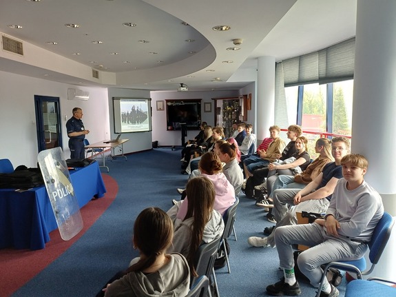 Licealiści siedzą na auli komendy policji i słuchaja prelekcji policjanta