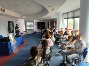 Licealiści siedzą na auli komendy policji i słuchaja prelekcji policjanta