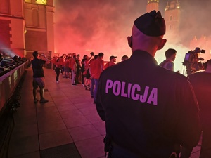 umundurowany policjant obserwuje w góry kibiców biorących udział w fecie. W tle widać dym oraz odpalone race