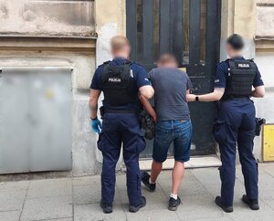 umundurowni policjanci prowadzą zatrzymanego mężczynę do budynku komisaraitu. Mężczyzna posiada kajdanki założone na ręce trzymane z tyłu