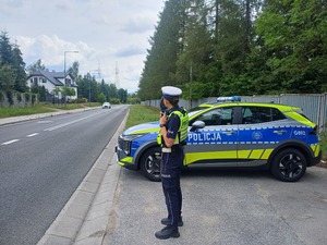 Policjantka stoi obok radiowozu na poboczu drogi i mierzy prędkość jadacym  samochodom.